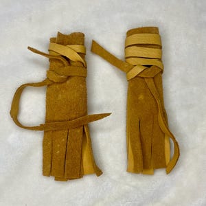 Op de afbeelding: Twee handgemaakte, bruinleren kwasten. Elke kwast is omwikkeld met een bijpassende leren strip, met lange, gefranjerde onderkanten. De kwasten worden gepresenteerd op een witte ondergrond.