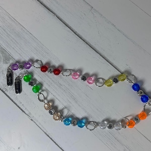 May include: A colorful beaded necklace with a variety of beads in different colors, including purple, red, green, pink, yellow, blue, and orange. The beads are strung together with silver-colored rings. The necklace also has two silver-colored rectangular charms.