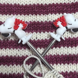 May include: Two metal knitting needles crossed over each other, each featuring a white Snoopy character topper holding a cluster of red hearts. The needles are positioned against a background of white and dark magenta striped knitted fabric.