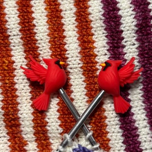Puede incluir: Dos marcadores de punto de p&aacute;jaro cardenal rojo con agujas plateadas cruzadas sobre una pieza de punto. El tejido tiene rayas verticales blancas, naranjas y moradas. Los cardenales tienen ojos negros y picos amarillos.
