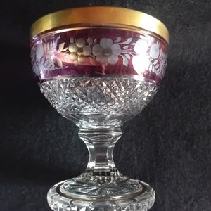 Può includere: Un calice di vetro trasparente con un designo in cristallo tagliato. La coppa del calice è decorata con una fascia di vetro viola e un bordo dorato. Il calice ha un motivo floreale inciso bianco.