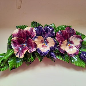 Peut inclure: Composition florale en céramique avec trois pensées aux tons de violet, crème et bordeaux, entourées de feuilles vertes brillantes. La pièce décorative est probablement destinée à être fixée au mur.