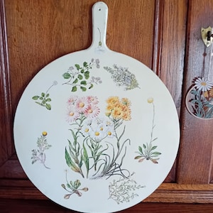 Può includere: Tagliere rotondo in legno bianco sporco con manico, decorato con illustrazioni botaniche di vari fiori e verde. I fiori sono in tonalità rosa, gialle e bianche, con foglie e steli verdi.