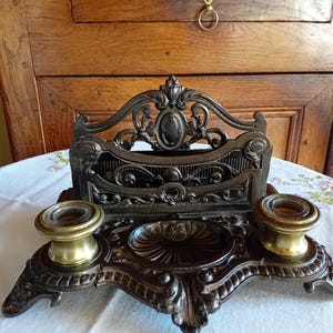 May include: A dark metal desk organiser featuring an ornate back panel with scrollwork, a central circular pen tray, and two brass-coloured inkwell holders with glass inserts. The base has decorative moulded edges and four small feet.