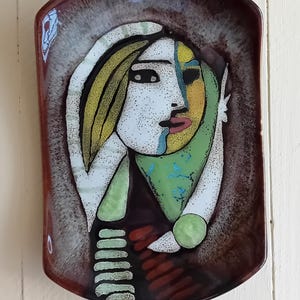 May include: A ceramic wall plate with a stylized portrait of a woman with yellow hair, green and yellow clothing, and a white background. The plate has a brown, speckled glaze.