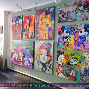 May include: A collection of vibrant pop art canvases displayed on a wall, featuring iconic characters like Mickey Mouse, Donald Duck, the Joker, and Marilyn Monroe, alongside stylized interpretations of famous paintings with graffiti-style paint splatters.