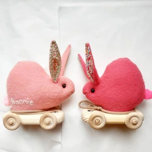 May include: Two pink plush bunny toys with floral-patterned ears and black eyes. Each bunny sits on a wooden cart with four wheels and a beige rope. One bunny has the name "Bonnie" embroidered on its side.