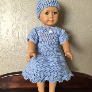 May include: A doll wearing a light blue crochet dress and matching hat. The dress has short sleeves and a scalloped hem. A small white snowflake adorns the chest. The doll is wearing red shoes.