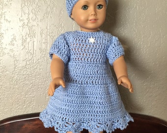 May include: A doll wearing a light blue crochet dress and matching hat. The dress has short sleeves and a scalloped hem. A small white snowflake adorns the chest. The doll is wearing red shoes.