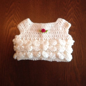 May include: White crocheted sleeveless top with a pink flower embellishment.