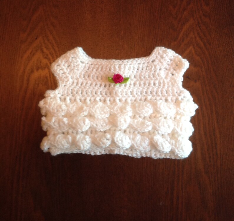 May include: White crocheted sleeveless top with a pink flower embellishment.