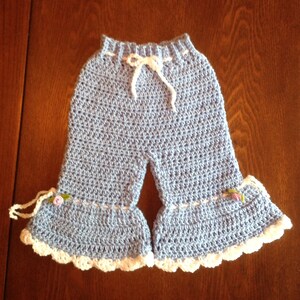 May include: Light blue crocheted baby pants with white trim and a white drawstring. The pants have a ruffled hem and small flower embellishments.