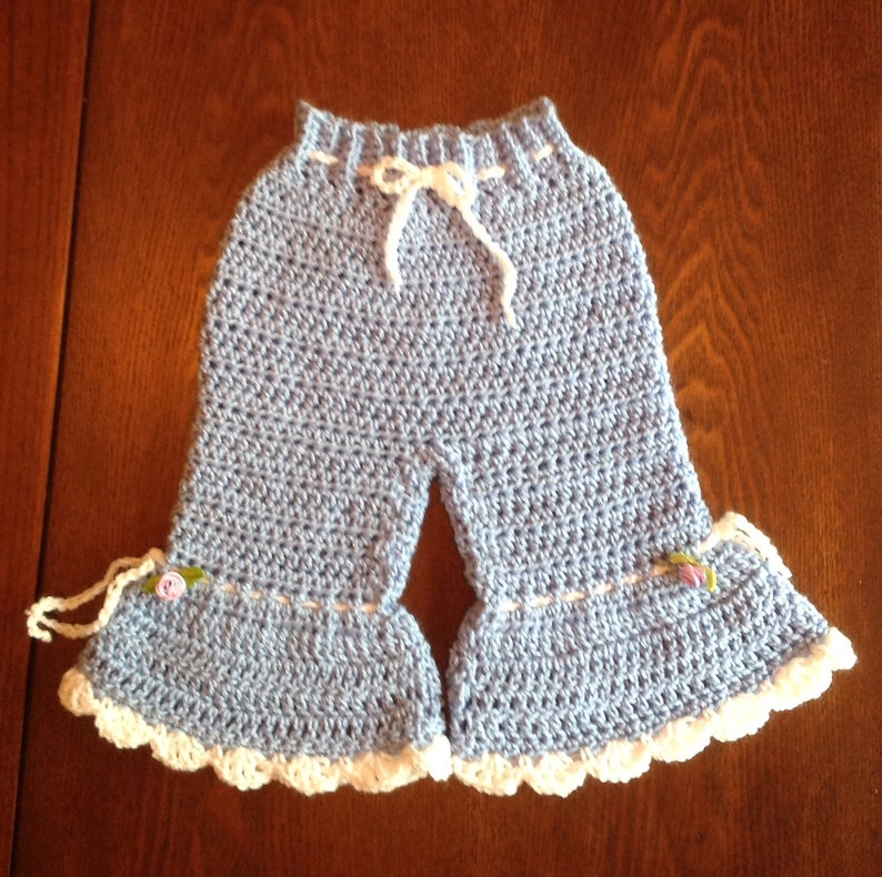 May include: Light blue crocheted baby pants with white trim and a white drawstring. The pants have a ruffled hem and small flower embellishments.