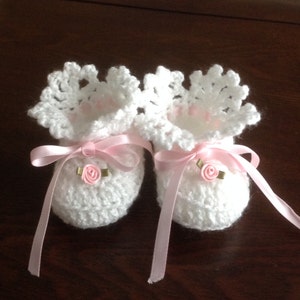 Pode incluir: Sapatinhos de bebé de crochet branco com laços de fita de cetim rosa e enfeites de rosa de tecido rosa.