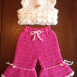 May include: A crocheted baby outfit with a white sleeveless top and pink pants. The top has a small pink flower embellishment. The pants have a white crocheted trim and a white bow at the waist.