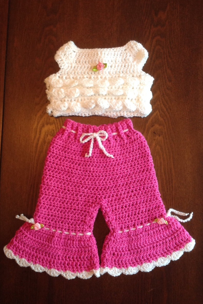 May include: A crocheted baby outfit with a white sleeveless top and pink pants. The top has a small pink flower embellishment. The pants have a white crocheted trim and a white bow at the waist.