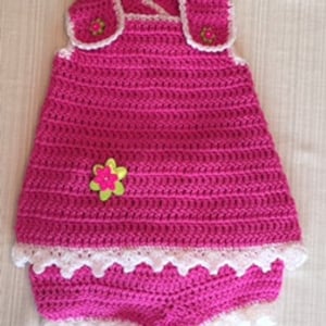 Pode incluir: Um vestido de bebé de crochet rosa com uma renda branca e um par de calções de crochet a condizer. O vestido tem um adorno de flor na frente.