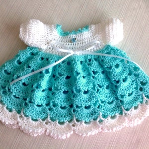 Può includere: Un vestitino da neonato all'uncinetto con un corpetto bianco e una gonna turchese. Il vestito ha un nastro bianco legato in vita.