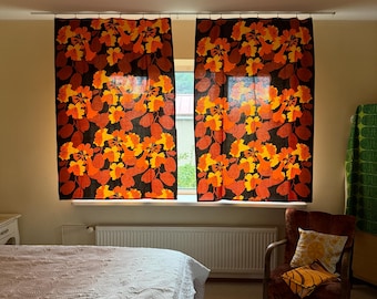 May include: Two window curtains featuring a bold floral pattern in shades of orange, yellow, and dark brown. The fabric displays clusters of stylized flowers and rounded leaves, hanging from a simple metal rod over a window.