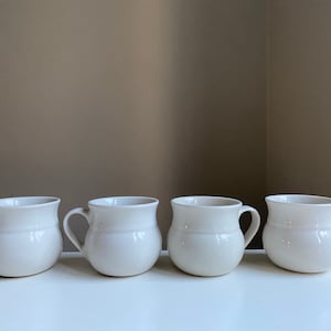 May include: Four white ceramic mugs with rounded bottoms and handles. The mugs are arranged in a row on a white surface.