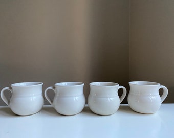 May include: Four white ceramic mugs with rounded bottoms and handles. The mugs are arranged in a row on a white surface.