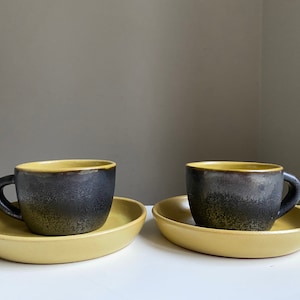 Puede incluir: Dos tazas de café de cerámica con platillos amarillos. Las tazas son negras con un borde amarillo y un esmalte moteado.
