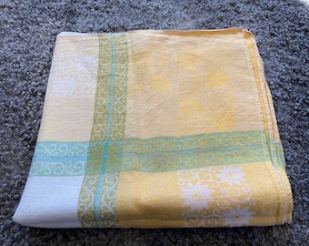 May include: A yellow and white tablecloth with a green and blue floral pattern. The tablecloth is folded in half and has a textured surface.