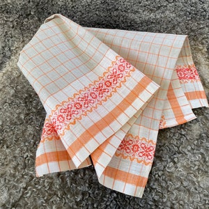 May include: Two white linen tea towels with orange and red embroidered borders. The towels are folded and displayed on a grey fluffy surface.