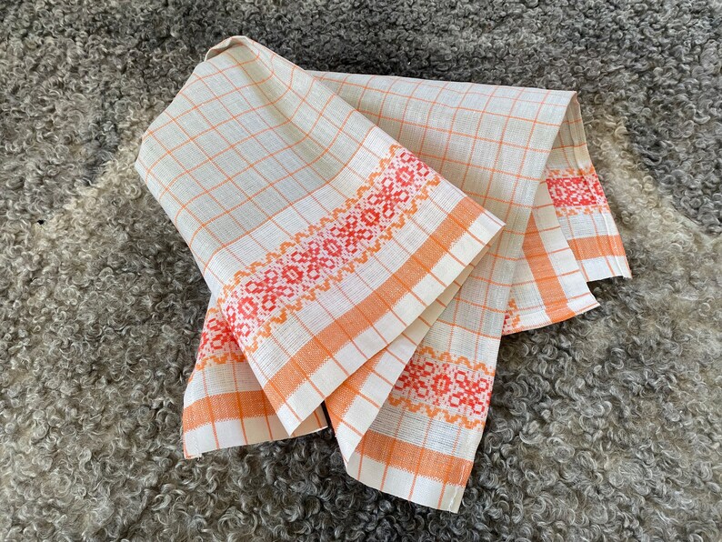 May include: Two white linen tea towels with orange and red embroidered borders. The towels are folded and displayed on a grey fluffy surface.