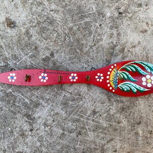 Può includere: Un cucchiaio di legno rosso con un motivo floreale dipinto e il testo "Buvas Brenna". Il cucchiaio ha cinque ganci di metallo sul retro.