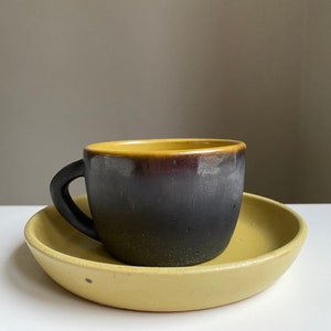 Puede incluir: Un juego de taza y platillo de café de cerámica amarilla y negra. La taza tiene un esmalte marrón oscuro en el exterior y un esmalte amarillo en el interior. El platillo es de color amarillo sólido.