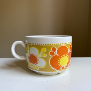 Puede incluir: Una taza de té de cerámica blanca con un patrón floral en tonos naranja, amarillo y marrón. La taza tiene una forma redondeada y un asa.