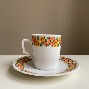 May include: A white ceramic coffee cup and saucer with a floral pattern in shades of orange, yellow, and green. The cup has a handle and the saucer has a scalloped edge.