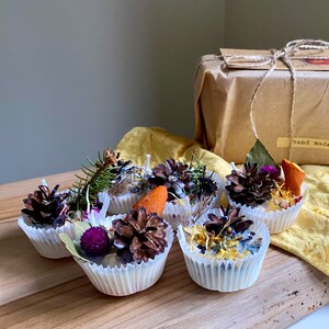 May include: Six small white candles with natural elements embedded in the wax. The candles are in white paper cupcake liners. The candles are arranged on a wooden surface. The candles are decorated with pine cones, dried flowers, and other natural elements. The candles are labeled "hand made".