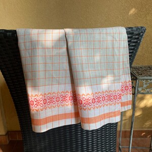 May include: Two white linen tea towels with orange and red checkered patterns and a decorative orange and red border. The towels are hanging on a black wicker chair.