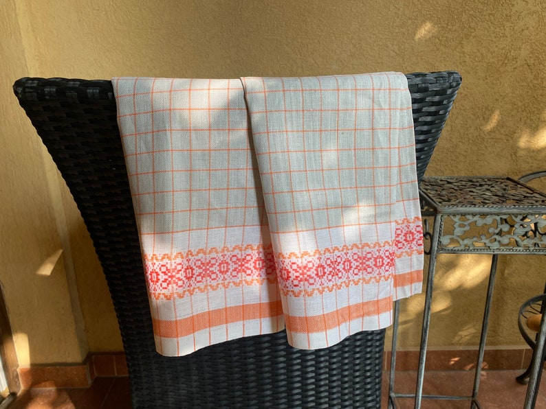 May include: Two white linen tea towels with orange and red checkered patterns and a decorative orange and red border. The towels are hanging on a black wicker chair.