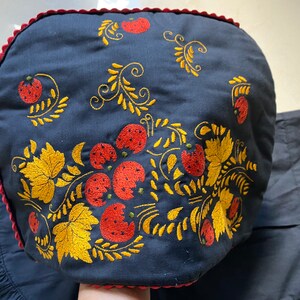 Può includere: Un cappello blu navy con ricami di fragole e foglie dorate. Il cappello ha un bordo rosso e un bordo a balze. Il ricamo presenta fragole rosse e foglie dorate.