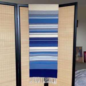 Könnte beinhalten: Ein gewebtes Textil mit Streifenmuster in Blau-, Weiß- und Grautönen. Das Textil hat einen Fransenrand am unteren Rand und wird vertikal angezeigt. Das Design weist horizontale Streifen unterschiedlicher Breite auf.