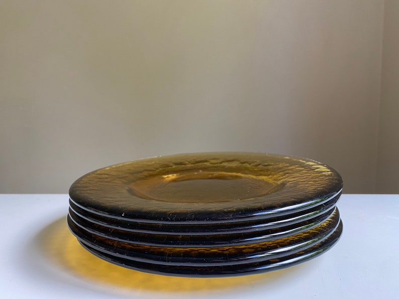 May include: Four amber glass dinner plates stacked on top of each other. The plates have a textured, rippled surface.