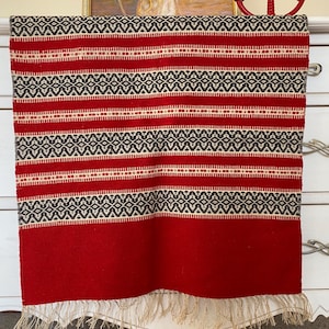 May include: Red and white woven table runner with a black geometric pattern. The runner has a fringe on the bottom edge.