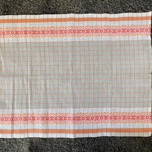 May include: A white kitchen towel with orange and red checkered pattern and a woven border with orange and red floral designs.