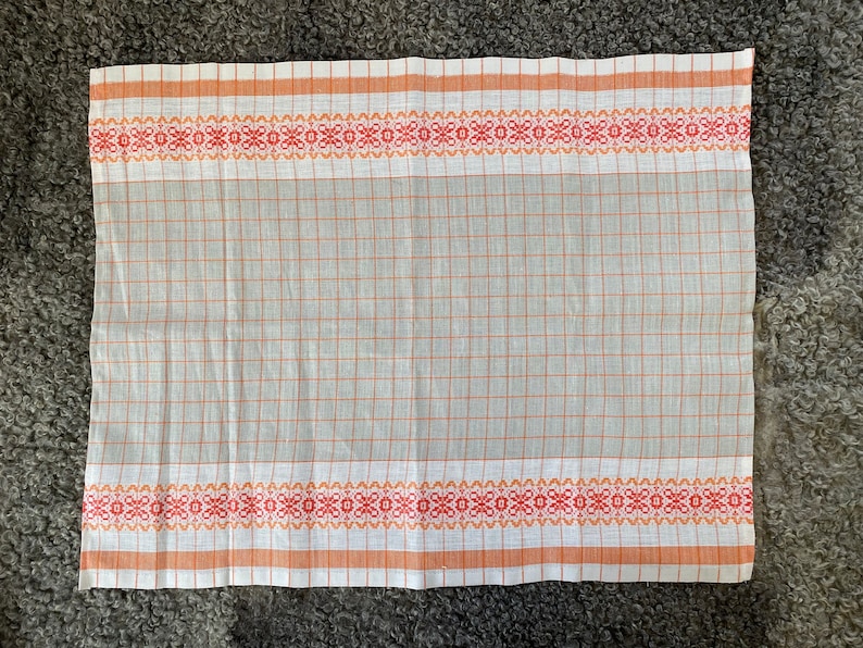 May include: A white kitchen towel with orange and red checkered pattern and a woven border with orange and red floral designs.