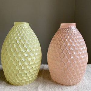 May include: Two decorative vases with a honeycomb pattern. One vase is a pale yellow-green, and the other is a soft pink. Both have a rounded, bulbous shape and a textured surface. The vases are displayed on a neutral-colored surface.