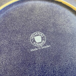 May include: Close-up of a blue ceramic plate with a white logo that reads "Hogan&auml;s Keramik" and "Made in Sweden".