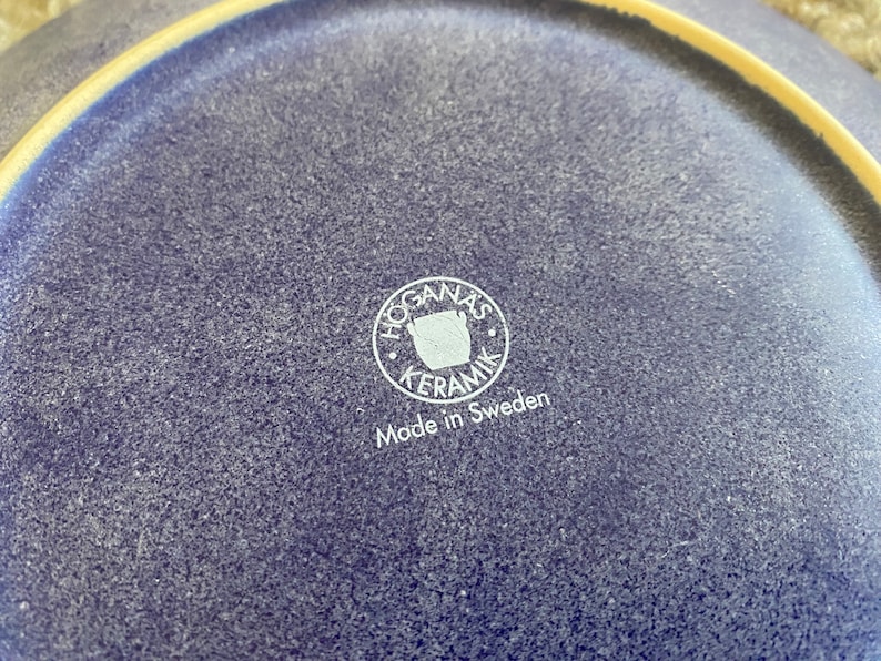 May include: Close-up of a blue ceramic plate with a white logo that reads "Hogan&auml;s Keramik" and "Made in Sweden".