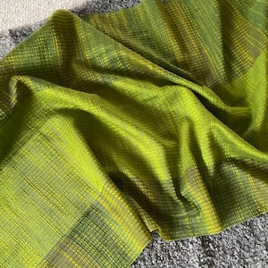 May include: A vibrant green scarf with a textured weave. The scarf features shades of lime and olive green, creating a gradient effect. The fabric is draped, showcasing its soft texture and potential for warmth and style.
