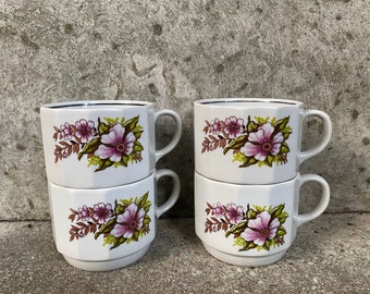 Op de afbeelding: Set van vier witte keramische mokken met bloemenmotieven. Elke mok heeft een roze en bruin bloemenmotief met groene bladeren. De mokken zijn in paren gestapeld.