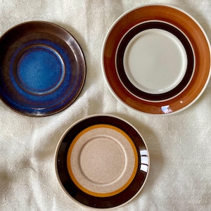 May include: Three ceramic saucer plates with different patterns. The first plate has a dark brown rim with a blue centre. The second plate has a brown rim with a white centre and brown and orange concentric circles. The third plate has a brown rim with a beige centre and brown and orange concentric circles.