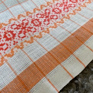 May include: A white linen tea towel with a red and orange embroidered border. The border features a geometric pattern.