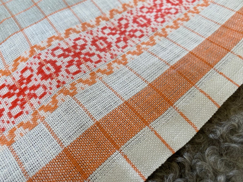 May include: A white linen tea towel with a red and orange embroidered border. The border features a geometric pattern.